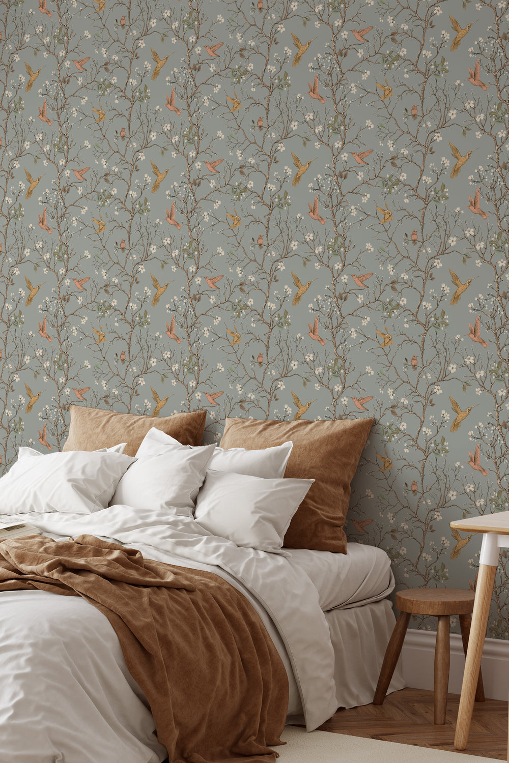 Sage gray hummingbird wallpaper with delicate flowering branches and colorful birds featured as a bedroom accent wall behind a cozy bed.