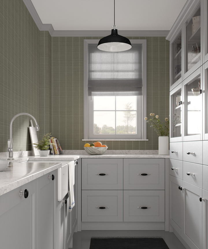 Sage gray Varrone canvas wallpaper with a subtle windowpane grid pattern covering the walls of a classic kitchen with white cabinetry, marble countertops, and a black pendant light.