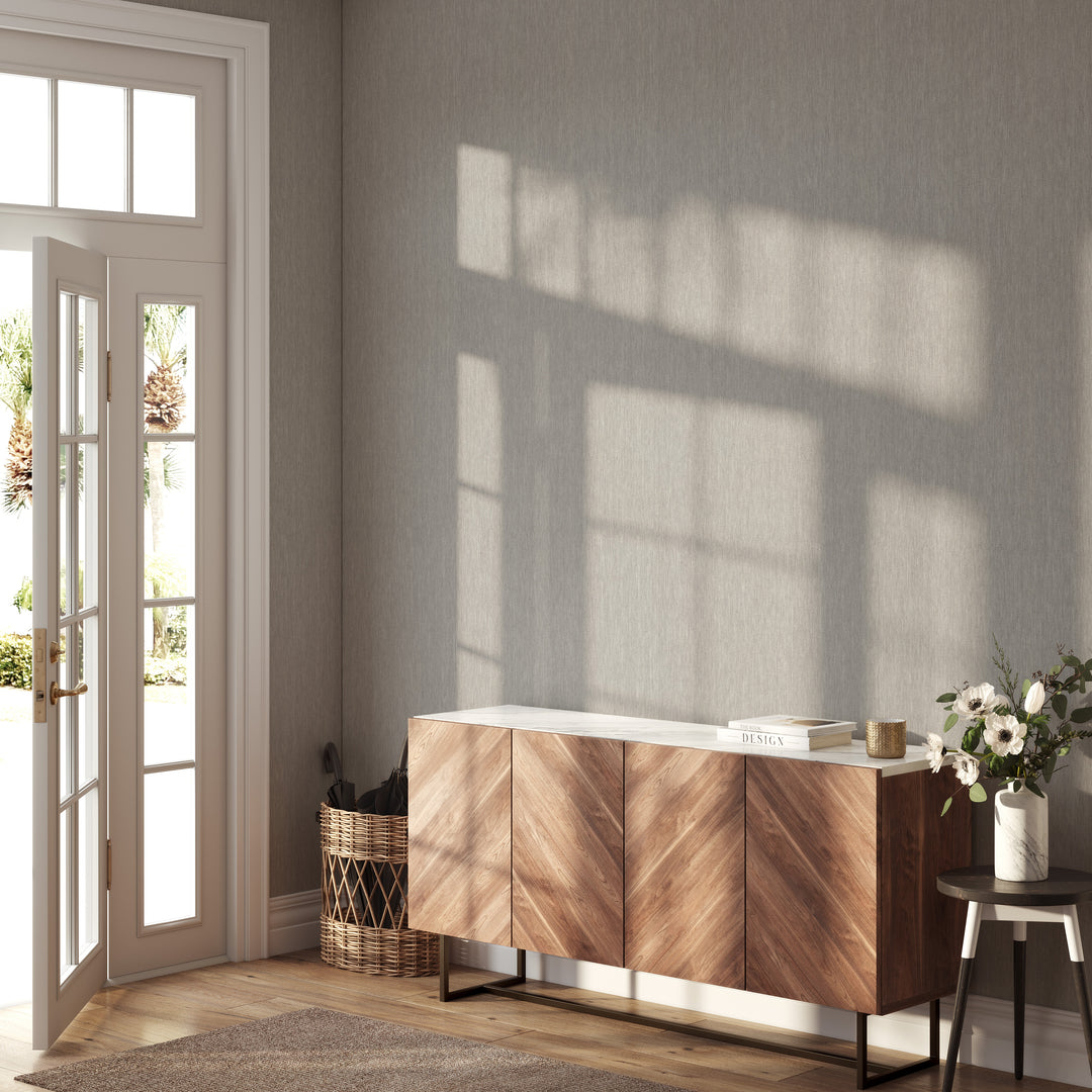 Light greige faux linen wallpaper with a subtle woven texture covering an entryway wall, paired with a warm wood sideboard, natural wood flooring, and soft sunlight streaming through an open door.