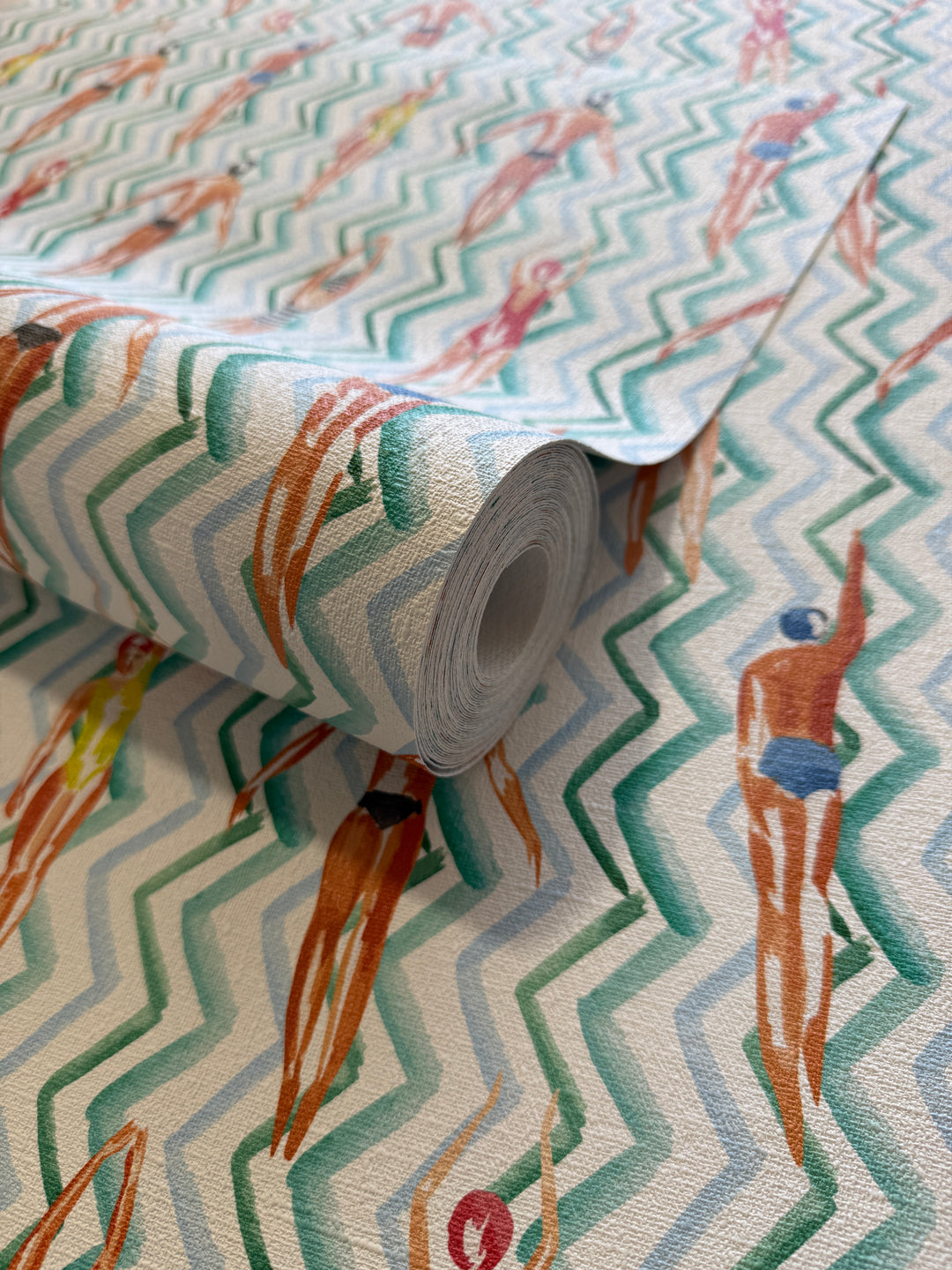 A close-up of a rolled sheet of wallpaper or fabric with a repeating chevron pattern in teal, mint, and cream, overlaid with illustrated figures in swimwear and beachwear.